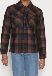 Brązowo-czarna flanelowa kurtka w kratę z kołnierzem, dwoma kieszeniami na piersi i zapięciem na guziki. Mieszanka bawełny o miękkiej teksturze.