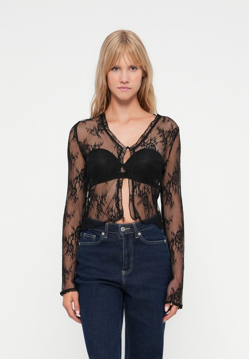 Svart blonde topp med lange ermer, dyp V-hals og en frontknute; gjennomsiktig stoff viser en svart bh under. Kombinert med mørke jeans.
