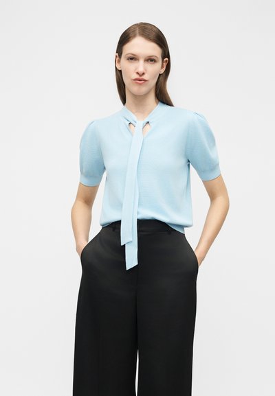 Γυναίκα που φοράει ανοιχτό μπλε κοντομάνικο τοπ με δέσιμο στο λαιμό και μαύρο παντελόνι ψηλόμεσο με φαρδιά πόδια, στέκεται με τα χέρια στις τσέπες.