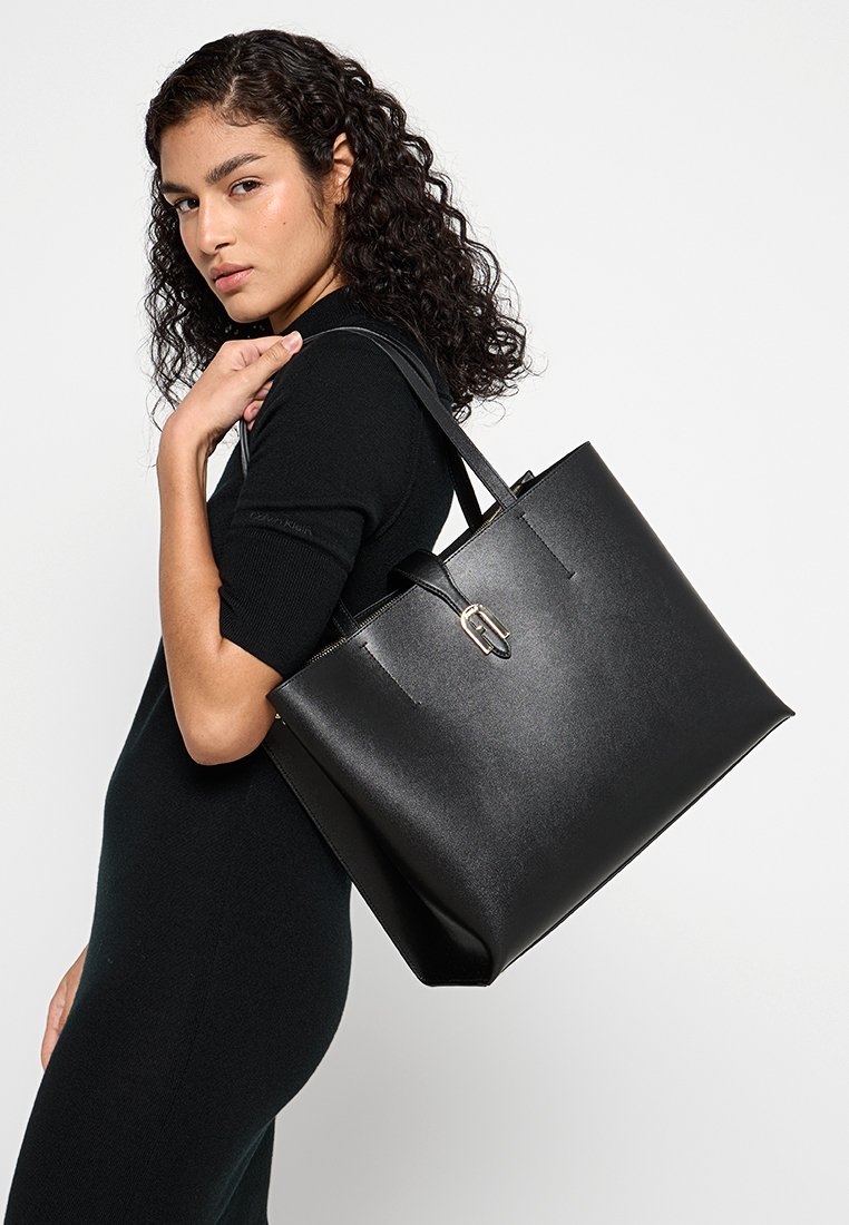 Femme aux cheveux bouclés portant une robe noire, tenant un grand sac en cuir noir sur son épaule, devant un fond uni.