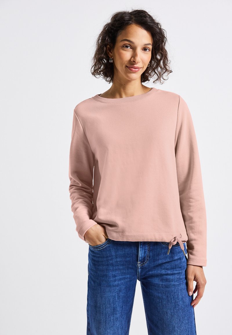 Ljusrosa sweatshirt med rundad halsringning, långa ärmar och dragsko i nederkanten. I kombination med blå denimjeans. Slät textur.