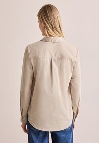 Blusa larga de manga larga color beige claro con cuello, pliegue en la parte posterior y puños abotonados. La tela tiene una textura suave y se combina con unos jeans azules.