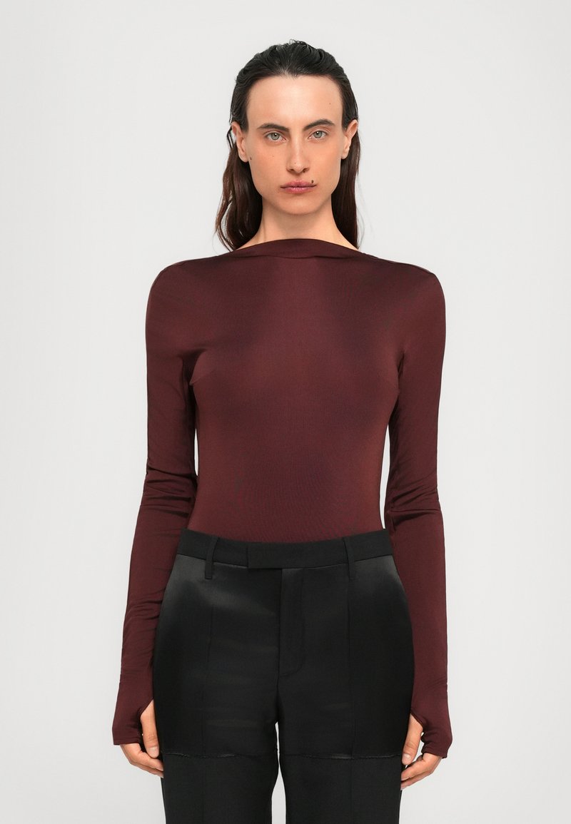 Woman with long dark hair wearing a fitted maroon long-sleeve top and high-waisted black pants against a plain background.