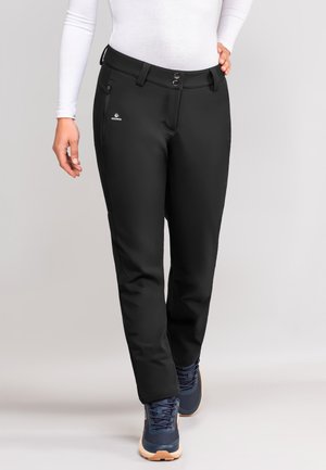 Zwarte outdoorbroek met een fitted ontwerp, twee zijzakken, een ritszak aan de zijkant en een logo aan de linkerkant. gedragen met marineblauwe schoenen.