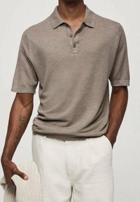 Homme portant un polo texturé taupe à manches courtes et un pantalon beige clair, tenant un pull beige tricoté dans la main gauche.