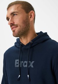 Felpa blu navy realizzata in tessuto morbido, con cappuccio con cordino e un grande logo "Brax" in una finitura opaca a contrasto.