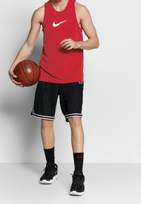 Débardeur rouge sans manches Nike, short noir avec bordure blanche, baskets noires, tenant un ballon de basketball, debout devant un fond neutre.