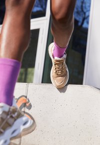 Persona che indossa sneaker color sabbia e calzini viola mentre sale su un barriera in cemento decorata con graffiti, all'aperto vicino all'ingresso di un edificio.