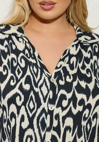 Schwarz-weiß gemusterte Hemdbluse mit Kragen, aus strukturiertem Stoff und abstrakten Motiven, die an Wirbel und Diamanten erinnern.