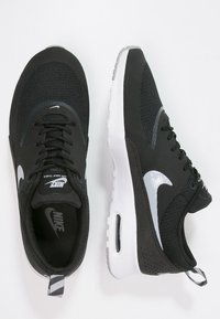 Nike Air Max Thea-skor har en svart mesh-övre, vit gummisula och grå detaljer, med en rund tå och snörningsdesign.