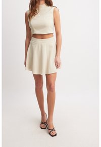 Vaaleankerman värinen kahden osan asu, joka koostuu lyhyestä hihattomasta tunikasta ja leveälahkeisesta mini-hameesta, valmistettu teksturoidusta kankaasta, ja asusteena mustat nyöritossut.