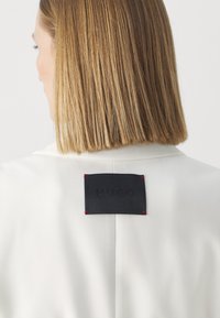 Biała marynarka z czarną metką z wytłoczonym napisem "HUGO", zaakcentowana czerwonym przeszyciem przy kołnierzu. Gładka faktura i dopasowany krój.