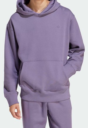 Mann trägt passenden lavendelfarbenen Hoodie und Jogginghose, eine Hand in der Hoodie-Tasche, Oberkörper und obere Beine sichtbar.