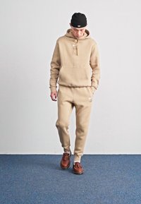 Beige Nike hettegenser med kengurulomme og snøring, sammen med matchende joggebukser. Brune sko med teksturert overflate. Enkelt design.