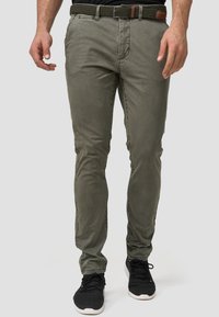 Pantalones de algodón verdes ajustados con bolsillos frontales y un cinturón tejido que presenta un acento de cuero marrón. Combinados con zapatos deportivos negros.