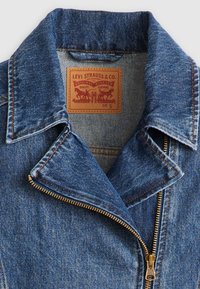 Denim takki sinisessä värissä, terävä kaulus ja kultainen vetskari. Siinä on ruskea etiketti, jossa on ompeluyksityiskohtia ja kokotagi sisällä.