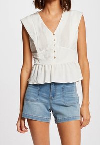 Top peplum blanco de lino con un profundo escote en V y mangas cortas, con botones dorados en la parte delantera. Combinado con pantalones cortos de mezclilla.