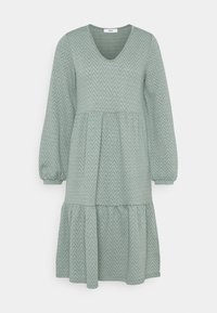 Robe verte texturée avec un décolleté en V, de longues manches bouffantes et un ourlet en volants. Présente un motif en chevrons sur l'ensemble du tissu.