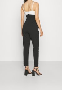 Femme de dos faisant face à la caméra, portant un pantalon noir taille haute, un haut sans manches blanc et des sandales à talons hauts noires.