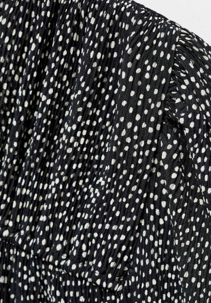 Tessuto nero con pois bianchi, caratterizzato da una texture pliss è. Il motivo è distribuito uniformemente, evidenziando il contrasto tra chiaro e scuro.