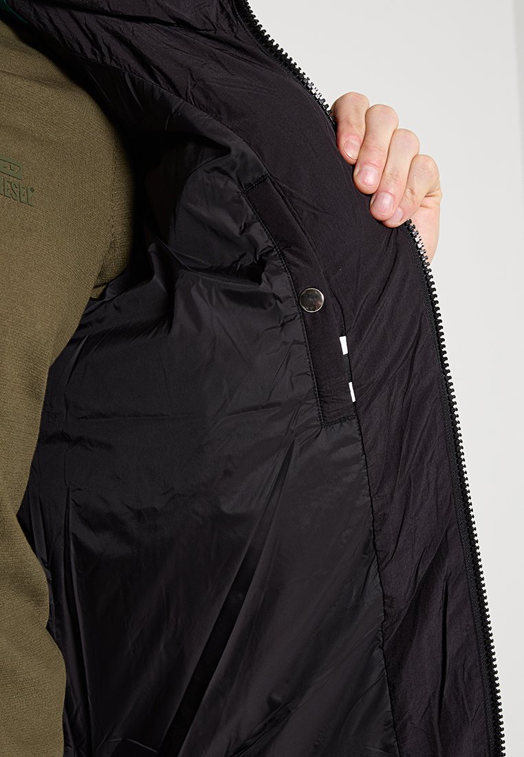 Mano che tiene aperto un giubbotto nero mostrando la tasca interna con bottone a pressione, indossato sopra una camicia verde oliva.