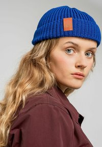 Niebieska dziana czapka beanie z teksturowanym wzorem i brązową skórzaną metką. Noszona z ciemnoczerwonym płaszczem, pokazująca zbliżenie z boku.