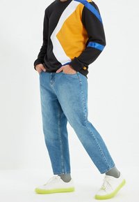 Musta collegepaita, jossa on keltaisia, valkoisia ja sinisiä geometrisia kuvioita, yhdistetty vaaleansinisiin farkkuihin ja valkoisiin lenkkareihin, joissa on limenvihreät pohjat.