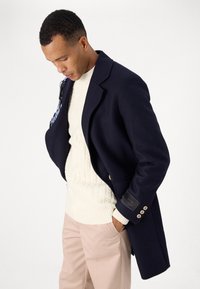 Palton din lână bleumarin, cu o croială elegantă, închidere cu două nasturi și un pulover cremos cu tricotaj tip cablu dedesubt. Pantaloni roz deschis completează ținuta.