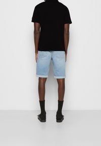 Mężczyzna ubrany w jasne, podwinięte szorty z denimu, czarną koszulkę polo, czarne skarpety i czarne sandały, stoi twarzą do białej, gładkiej ściany.