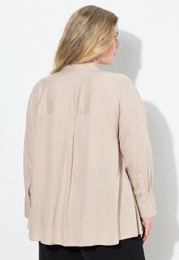 Blusa beige a maniche lunghe realizzata in tessuto morbido; presenta un dettaglio sul retro con yoke e polsini abbottonati, con una vestibilità rilassata e una silhouette drappeggiata.