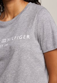 Person iført en lys grå Tommy Hilfiger T-shirt med korteærmer og trykt mærke navn tekst på brystet.