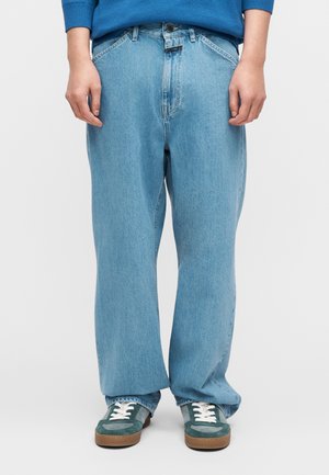 Hellblaue, weit geschnittene Jeans mit hoher Taille und kontrastierender Naht. Kombiniert mit grünen und braunen Sneakern mit weißen Schnürsenkeln.