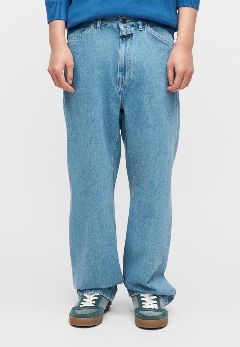 Ανοιχτό μπλε τζιν με φαρδιά μπατζάκια και ψηλή μέση, με αντίθετη ραφή. Φορώντας πράσινα και καφέ πάνινα παπούτσια με λευκά κορδόνια.