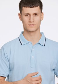 Jasnoniebieska koszulka polo z teksturowanej tkaniny, z trzema guzikami, granatowym, prążkowanym kołnierzykiem i subtelnym logo na klatce piersiowej.