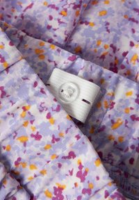 Tissu floral en violet pastel, décoré de petits groupes de taches roses, jaunes et blanches. Comprend un détail avec un bouton blanc et une sangle.