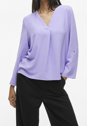 Vrouw draagt een losse lavendelkleurige V-hals blouse met gestructureerde stof en lange mouwen, gecombineerd met zwarte broek, staand met één hand in de zak.