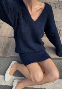 Mörkblå stickad tröja med V-ringning och långa ärmar; mjuk textur, något oversized passform, ihop med vita platta skor.