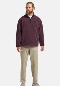 Maroon fleece pullover with a zip neck, black accents, and a logo patch. Pair of light tan pants; individual stands on neutral background.