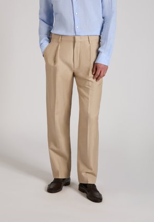 Mężczyzna ubrany w beżowe plisowane spodnie, jasno niebieską koszulę na guziki włożoną do spodni oraz ciemnobrązowe skórzane buty, stojący z jedną ręką w kieszeni.