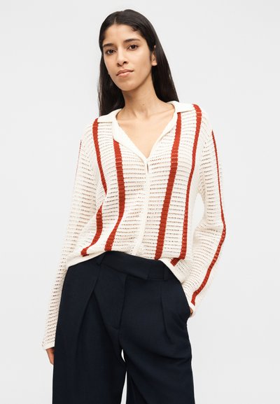 Γυναίκα που φοράει λευκό πλεκτό πουλόβερ με κάθετες κόκκινες ρίγες και μαύρο παντελόνι με φαρδιά πόδια, στέκεται με το ένα χέρι στην τσέπη, ουδέτερο φόντο.