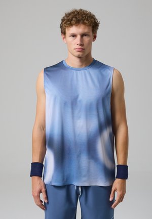 Giovane uomo con capelli ricci che indossa una maglietta sportiva blu senza maniche, pantaloncini abbinati e fasce scure ai polsi su uno sfondo grigio chiaro.