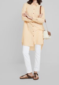 Lichtoranje, knoopshirt met kraag en verticale strepen, gecombineerd met witte rechte broek en bruine flip-flops. Kleine geweven handtas.