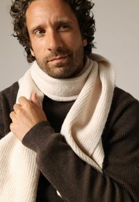 Écharpe crème côtelée drapée autour du cou, en tissu doux. Pull texturé marron foncé en dessous, manches longues visibles.