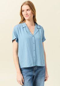 Camisa de manga corta azul claro con cuello, botones frontales y un patrón texturizado. Combinada con jeans de mezclilla azul claro.