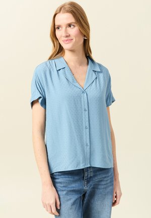 Camicetta azzurra a maniche corte con colletto, bottoni frontali e motivo testurizzato. Indossata con jeans in denim azzurro.