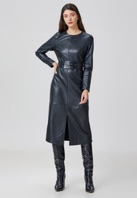 Robe en cuir noir à manches longues, silhouette ajustée, taille ceinturée, fente hauteur genoux et texture lisse. Associée à des bottes hautes assorties.