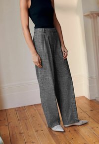 Donna che indossa pantaloni grigi a vita alta e a gamba larga, top senza maniche nero, scarpe bianche con punta, in piedi su un pavimento in legno all'interno.