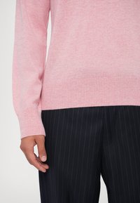 Svijetloružičasti pleteni pulover s rebrastim manžetama i rubom, u kombinaciji s tamnim prugastim hlačama. Fokus na teksturu i suptilne varijacije boje.