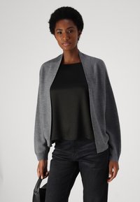Cárdigan gris hecho de tela texturizada, con abertura frontal, corte relajado y mangas anchas, usado sobre una camiseta negra y combinado con jeans oscuros.