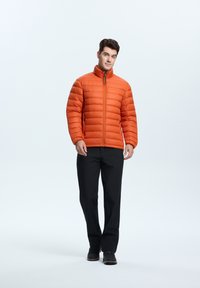 Orange polstret jakke med høj krave, lynlåslukning og sorte detaljer, båret over sorte bukser og udendørssko, mod en ensfarvet baggrund.
